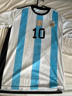 Argentina fotbollströja Messi #10 - Säljer en officiell Argentina landslagströja från Adidas med Messis namn och nummer 10. Tröjan är vit med ljusblå ränder, svarta detaljer och AFA-emblem på bröstet. Perfekt för fotbollsfans som vill stötta sitt lag och idol.