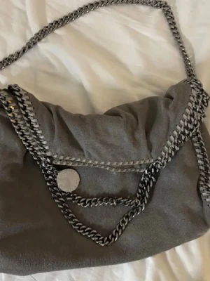 Stella McCartney väska - Säljer min fina gråa axelbands/handväska från Stella McCartney i modellen Falabella i mellan storlek. Rymlig insida - storleken är perfekt för att ha en dator i och lite annat - med rosa foder. Väskan har silvriga kedjedetaljer. Ordinarie pris ca 12.000 kr. En av sidorna har ett minimalt hål som knappt syns vid användning (se sista bild), därav detta superbra pris! Vid fler bilder eller äkthetsbevis är det bara att kontakta! 