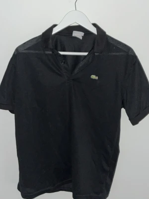 Svart pikétröja från Lacoste - Klassisk svart pikétröja från Lacoste med korta ärmar och den ikoniska krokodilloggan på bröstet. Tröjan har krage och knäppning framtill. Perfekt för en clean och sportig look. Pris kan diskuteras 