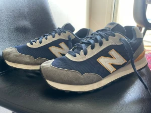 New Balance 515 blå sneakers - Snygga New Balance 515 sneakers i marinblått mesh och mocka med grå och vita detaljer samt orange kant runt loggan. Klassisk retrostil med vita sulor och mörkblå snören. Perfekta för dig som gillar streetwear och sportig look.