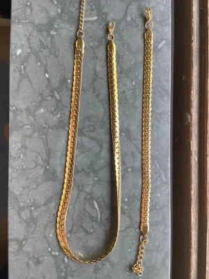 Guldigt platt halsband - Snyggt halsband i guldfärgad metall med platt, glänsande kedja och klassisk karbinhake. Kedjan har en modern, stilren design som passar perfekt till olika looks. Inga synliga märken eller logotyper på halsbandet.