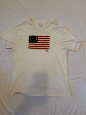 Vit t-shirt med amerikansk flagga RL - Vit t-shirt från Polo Ralph Lauren med broderad amerikansk flagga på bröstet och röda initialer 'R.L.' under flaggan. Klassisk passform och rund hals. Perfekt för dig som gillar stilrena plagg med en twist.