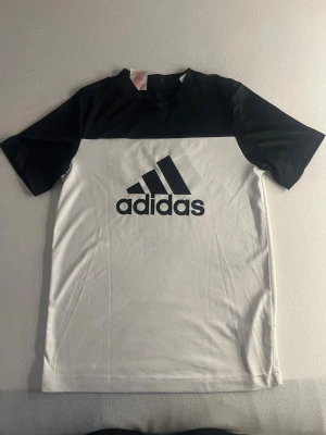 Svartvit Adidas t-shirt med logga - Snygg svartvit t-shirt från Adidas med stor logga på bröstet. T-shirten har korta ärmar och rund hals. Tillverkad i mjuk bomull som känns skön mot huden. Perfekt för en sportig och avslappnad stil.