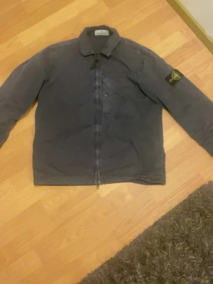 Mörkgrå overshirt från Stone Island - Säljer en mörkgrå overshirt från Stone Island med dragkedja framtill och klassisk krage. Ikonisk Stone Island-patch på vänster ärm. Overshirten är tillverkad i ett slitstarkt bomullstyg och har en clean, rak passform som funkar till många olika stilar.