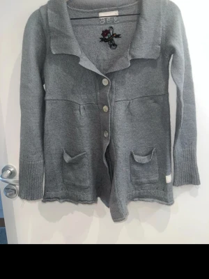 Grå stickad kofta med fickor - Mysig grå stickad kofta med fyra knappar framtill och två små fickor. Koftan har en bred krage och en broderad detalj med ros vid halsen. Perfekt att slänga på sig över en t-shirt eller topp.