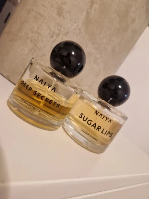 NAIYA Deep Secrets & Sugar Lips parfym - Två parfymer från NAIYA: Deep Secrets och Sugar Lips. Båda kommer i runda glasflaskor med svart klotformad kork och tydlig text på framsidan. Vätskan är ljusgul och flaskorna har en stilren, modern design. ( Se mängd kvar) 