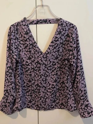 Lila blommig blus Gina Tricot - Snygg blus från Gina Tricot i svart med lila blommönster. Blusen har djup V-ringning och långa ärmar. Materialet är lätt och flowy, perfekt för att lyfta din outfit med ett trendigt mönster.