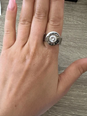 Silverfärgad ring med stor sten från LenMer - Säljer en chunky silverfärgad ring i metall med en stor, klar rund sten i mitten. Ringen har en bred design och text ingraverad runt stenen. Perfekt statement piece för dig som gillar att sticka ut. Ringen är i storlek 6 och aldrig använd! OBS: FRAKTAR INOM 24H!🩷