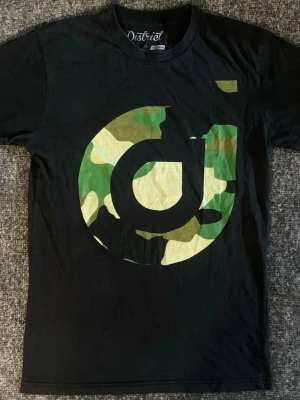 Svart t-shirt med camotryck från District - Svart t-shirt från District med stort grafiskt tryck i camomönster på bröstet. Klassisk rund halsringning och rak passform. Tillverkad i 100% bomull för en skön känsla och snygg streetstil.