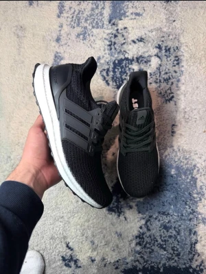 Svarta Adidas Ultraboost sneakers - Säljer ett par svarta oanvända Adidas Ultraboost sneakers med stickad ovandel och vita Boost-sulor. Skorna har svarta plastdetaljer på sidorna och klassiska tre ränder. Perfekta för dig som gillar stilrena och bekväma sneakers.