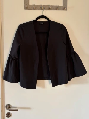 Svart kavaj med vida ärmar - Stilren svart kavaj med öppen framsida och vida trumpetärmar med rynkdetalj. Perfekt för att lyfta en enkel outfit och ge en trendig touch. Tillverkad i ett mjukt tyg som känns bekvämt mot huden.