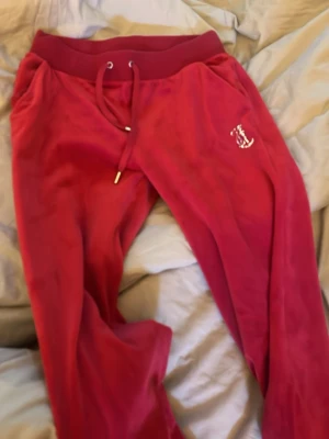 Röda Juicy Couture mjukisbyxor - Säljer ett par klassiska röda mjukisbyxor från Juicy Couture i mjuk velour. Byxorna har resårmidja med dragsko, fickor och glittrande Juicy-text baktill. Perfekta för en chill och trendig look.