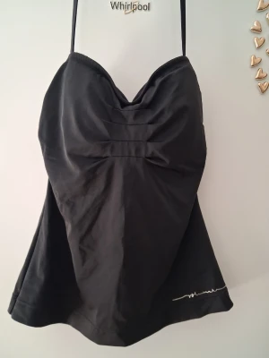 Svart tankini baddräkt med tunikaform - Snygg svart tankini baddräkt med tunikaform och smala axelband. Draperad byst och lätt utställd nederdel ger en feminin siluett. Materialet ser ut att vara stretchigt och snabbtorkande, perfekt för stranden eller poolen. Diskret vit detalj nertill.