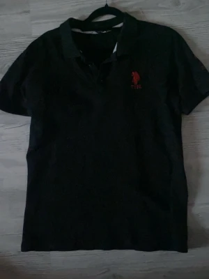 Svart pikétröja med röd logga - Snygg svart pikétröja med klassisk krage och knappar framtill. På bröstet finns en röd broderad logga som ger en sportig touch. Perfekt till jeans eller shorts för en avslappnad stil.