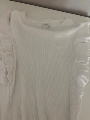 Vit ribbad topp med volang från Zara - Supersöt vit ribbad topp från Zara med volangdetaljer längs axlarna. Perfekt för dig som gillar en stilren look med en twist. Toppen är ärmlös och har en mjuk, stretchig känsla.