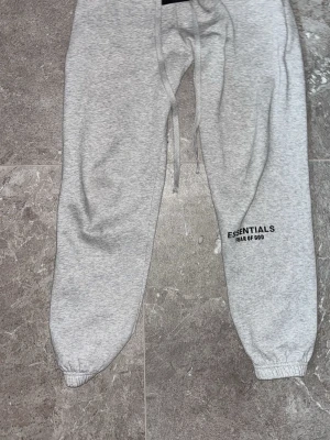 Grå mjukisbyxor Essentials Fear of God - Snygga grå mjukisbyxor från Essentials Fear of God med svart logga på benet. Byxorna har resår i benslut och midja samt dragsko för bästa passform. Perfekta för en chill och avslappnad stil.