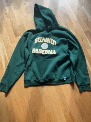 Russell athletic hoodie - Mörkgrön hoodie från Russell Athletic med stor gul och vit 'Belhaven Baseball'-text och basebollmotiv på bröstet. Klassisk känguruficka framtill och huva med dragsko. Tillverkad i mjuk bomullsblandning, perfekt för chill dagar eller sportig stil.