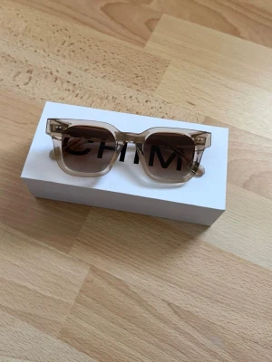 Beige Chimi 04 - Snygga solglasögon från CHIMI med transparent beige båge och fyrkantig form. Glasen är bruna med en lätt toning som ger en cool look. Perfekta för soliga dagar och ger en trendig vibe till din outfit.