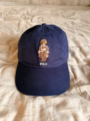 Marinblå Polo Ralph Lauren keps - Snygg marinblå keps från Polo Ralph Lauren med den klassiska björnen broderad framtill och justerbart spänne bak. Kepsen har böjd skärm och är tillverkad i bomull. Perfekt accessoar för en avslappnad och trendig look.