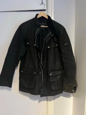 Svart vaxad jacka från Barbour - Klassisk svart vaxad jacka från Barbour International med quiltat innerfoder, flera stora fickor med tryckknappar och dragkedja framtill. Jackan har en ståkrage med manchesterdetalj och patch med Barbour-logga på ärmen. Perfekt för en cool och tidlös look.