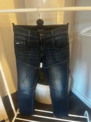 Mörkblå jeans med kontrastsömmar - Snygga mörkblå jeans med slitningar och kontrastsömmar. Modellen har fem fickor, guldfärgade nitar och detaljerade bakfickor. Jeansen har en modern passform och markerade sömmar över knäna för en cool look. W32 L32. Hör av er vid frågor!