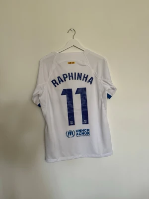 Vit FC Barcelona matchtröja Raphinha - Säljer en vit FC Barcelona matchtröja från Nike med Raphinha och nummer 11 på ryggen. Tröjan har klubbmärke och Nike-logga på bröstet, samt Spotify som sponsor. Ärmarna har blå och röda detaljer. Perfekt för fotbollsfans!
