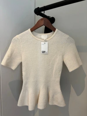 Vit ribbstickad topp med volangkant från hm - Supersöt vit ribbad topp med korta ärmar och rund hals. Toppen har en markerad midja och volangkant nedtill som ger en feminin siluett. Perfekt att styla med jeans eller kjol för en trendig look. Prislapp kvar!