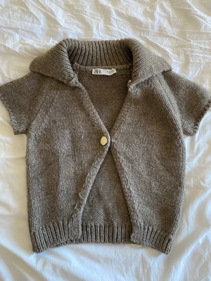 Kortärmad stickad kofta från Zara - Kortärmad stickad kofta från Zara i en mjuk beige färg. Koftan har en bred krage, en knapp framtill och ribbade detaljer vid ärmslut och nederkant. Perfekt att slänga över en topp för en chill look.