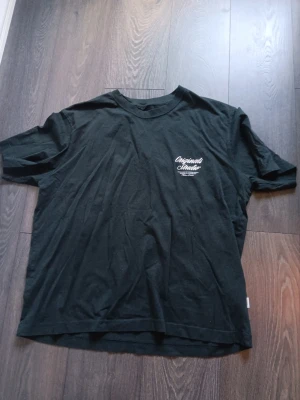 Mörk grön t-shirt med tryck - Säljer en  t-shirt med vit text på bröstet och stort tryck på ryggen där det står 'Originals Studio'. T-shirten har rund hals och korta ärmar. Perfekt för dig som gillar enkel men snygg streetwear. Använd 1 gång