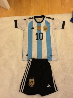 Argentina matchställ med nummer 10 - Säljer ett Argentina matchställ med klassiska blå och vita ränder. Tröjan har nummer 10 och AFA-emblem på bröstet samt svarta detaljer. Tillhörande svarta shorts med vita ränder och AFA-logga. Perfekt för fotbollsträning eller match. Pris kan diskuteras vid snabb och enkel affär!