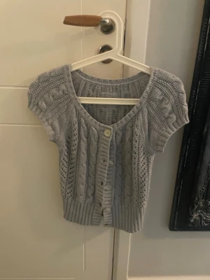 Söt grå kortärmad stickad kofta - Jätte gullig kortärmad stickad kofta i ljusgrått med kabelstickat mönster och dekorativa hålstickningar. Koftan har rund halsringning, ribbade kanter och knäppning framtill med fyra knappar. 
