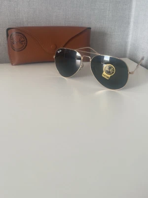 Ray-Ban Aviator solglasögon guld - Säljer ett par klassiska Ray-Ban Aviator solglasögon med guldfärgade metallbågar och mörka linser. Medföljer originalbrun läderfodral. Snygg och tidlös modell som passar till allt och ger en cool vibe.
