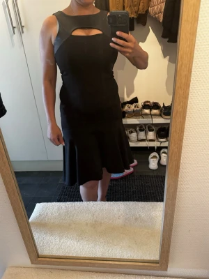 Svart klänning med cut out-detalj - Säljer en svart klänning med snygg cut out-detalj vid halsen och figurnära passform. Klänningen har ärmlös design och en utsvängd kjol som ger en feminin siluett. Perfekt för dig som vill ha en stilren och trendig look.