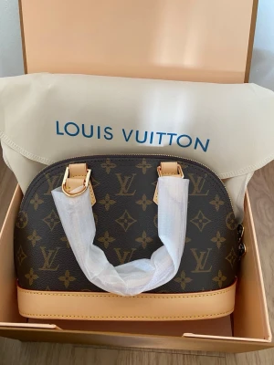 Louis Vuitton monogram handväska - Snygg handväska från Louis Vuitton i klassiskt brunt monogrammönster med ljusa läderdetaljer och guldfärgade metalldelar. Väskan har rundad form och två handtag. Perfekt för dig som gillar lyxiga accessoarer och tidlös design.