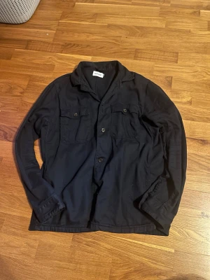 Mörkblå overshirt - Stilren mörkblå overshirt med två bröstfickor och knäppning framtill. Overshirten har klassisk krage och långa ärmar, perfekt att slänga på sig över en t-shirt. Materialet är mjukt och bekvämt, passar till många olika outfits.