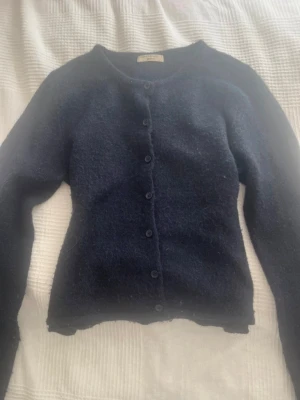 Mörkblå kofta från Gina Tricot - Mysig mörkblå kofta från Gina Tricot med rund halsringning och knappar framtill. Koftan är stickad i ett fluffigt material och har långa ärmar. Perfekt att slänga på sig när det är lite kyligare ute.