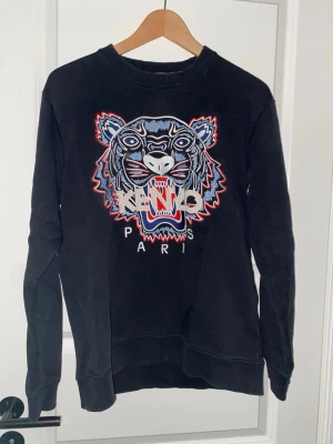 Svart sweatshirt från Kenzo med tiger - Svart sweatshirt från Kenzo med ikonisk broderad tiger i blått, vitt och rött framtill. Rund halsringning, långa ärmar och ribbade muddar. Stort Kenzo Paris-tryck under tigern. Perfekt statement-plagg för dig som gillar streetwear.