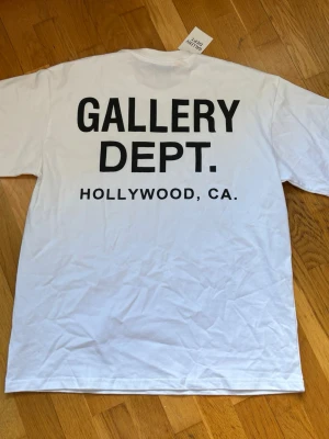 Vit t-shirt med tryck från Gallery Dept. - Vit t-shirt med stor svart text 'GALLERY DEPT. HOLLYWOOD, CA.' på ryggen. Klassisk passform och rund hals. Perfekt för dig som gillar streetwear och tydliga statement-plagg. Oversize baggy fit