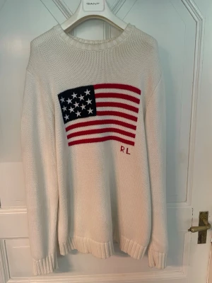 Vit stickad tröja med USA-flagga - Vit stickad tröja med en stor amerikansk flagga i rött, vitt och blått på bröstet samt bokstäverna RL broderat. Tröjan har rund halsringning och ribbade muddar vid ärmslut och nederkant. Tillverkad i bomull för en mjuk och bekväm känsla.