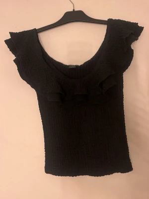Svart volangtopp från Lindex - Supersnygg svart topp från Lindex i smockat material med bred volang runt ringningen. Toppen är ärmlös och har en figurnära passform som framhäver siluetten. Perfekt att styla med jeans eller kjol för en trendig look.aldrig använd 