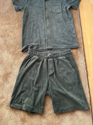 Mörkgrönt loungewear-set i frotté - Mysigt loungewear-set i mörkgrön frotté med kortärmad skjorta och matchande shorts. Skjortan har knappstängning och bröstficka, shortsen har resår i midjan och fickor både fram och bak. Perfekt för chill dagar hemma.