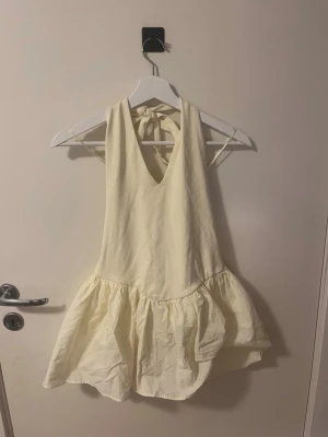 Ljusgul halterneck klänning - Supersöt ljusgul klänning med halterneck och knyt i nacken. Klänningen har en djup v-ringning fram och voluminös kjol. Tillverkad i ett lätt tyg som ger en luftig känsla, den är från Zara och är helt oanvänd med öppen rygg 