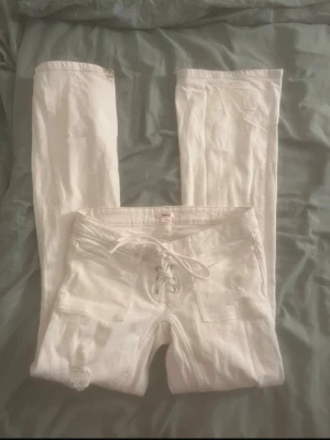 Vita snörade bootcut jeans - Säljer ett par vita jeans med bootcut-modell och snörning framtill i midjan. Jeansen har fickor både fram och bak samt en liten slitning vid ena benet. Materialet är denim med stretch för skön passform. Dom är lite smutsig där nere som man kan se i bild 3