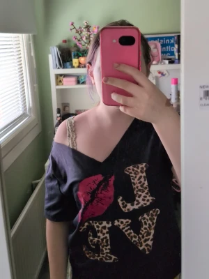 Svart croppad topp med leopardtryck - Svart croppad topp med v-ringning och korta ärmar. Framsidan har texten 'LOVE' där bokstäverna är i leopardmönster och ett stort rosa läppavtryck. Toppen har en avslappnad passform och är perfekt för en trendig och cool look.