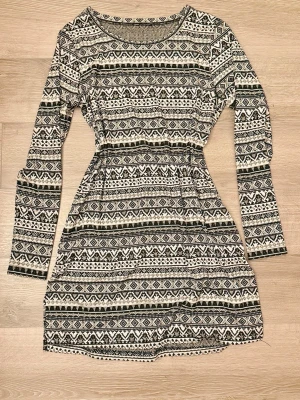 Svartvit mönstrad långärmad klänning L/40 - Kort klänning i svartvitt med grafiskt mönster. Långärmad modell med rund hals och markerad midja. Tillverkad i polyester, viskos och elastan för en mjuk och stretchig känsla. Storlek Large.