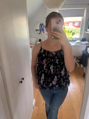 Blommig blå linnetopp - Säljer en blå linnetopp med blommigt mönster i rosa och vitt. Toppen har tunna axelband och en lös, flowy passform som känns perfekt för varma dagar. Materialet är lätt och luftigt, vilket gör den superbekväm att bära. Stlk 11/12 år