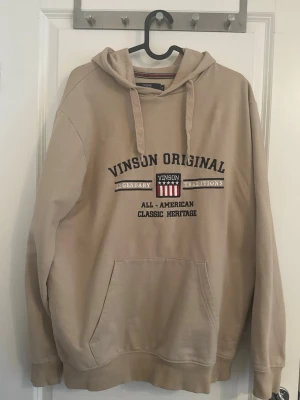 Beige hoodie från Vinson Original - Snygg beige hoodie från Vinson Original med tryck på bröstet och klassisk känguruficka. Huva med dragsko och mjukt material som känns skönt mot huden. Perfekt för en avslappnad och stilren look.
