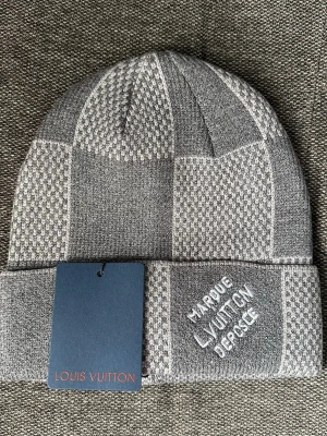 Grå mössa med rutmönster - Snygg grå mössa med rutigt mönster i olika nyanser av grått. Broderad text framtill. Tillverkad i mjukt stickat material som håller dig varm och stilren under alla dagar.