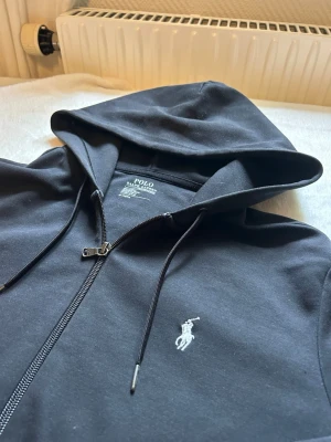 Svart hoodie från Polo Ralph Lauren - Svart hoodie med dragkedja från Polo Ralph Lauren. Klassisk design med vit broderad logga på bröstet, huva med dragsko och två fickor framtill. Tillverkad i mjukt bomullsmaterial som är skönt att bära.