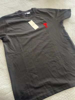 Svart t-shirt från Ami med rött hjärta - Svart t-shirt från Ami Alexandre Mattiussi med klassisk passform och rund hals. På bröstet finns ett broderat rött hjärta med bokstaven A. Tillverkad i mjuk bomull, perfekt för en stilren och trendig look.